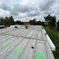 Budowa domu w toku. Widok na strop z prefabrykatów, zbrojenie czeka na beton. W tle drzewa i pochmurne niebo.