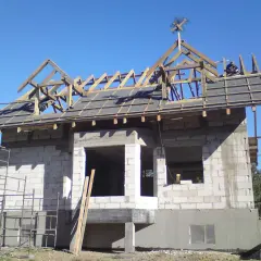 Na dachu domu w budowie, wśród drewnianej konstrukcji, widoczna jest tradycyjna wiecha.