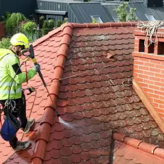 Pracownik w kasku i kamizelce odblaskowej wykonuje mycie dachu z użyciem myjki ciśnieniowej.