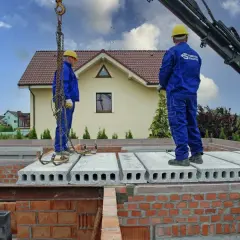 Dwóch pracowników w niebieskich kombinezonach montuje prefabrykowany strop. Dźwig podnosi kolejne płyty stropowe.