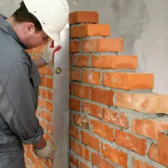 Murarz w kasku i rękawicach sprawdza pion muru z cegły, dbając o prawidłowe wymiary cegły i równość ścian.