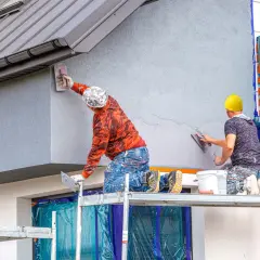 Dwóch budowlańców nakłada tynki mineralne na ścianę budynku, pracując na rusztowaniu.
