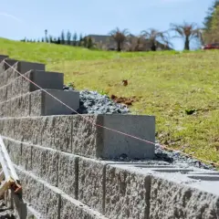 Budowa muru oporowego z bloczków betonowych na zboczu. Widać drabinę i sznurek do poziomowania.