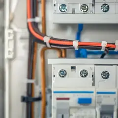 Zabezpieczona instalacja elektryczna w garażu: przewody w kolorach pomarańczowym i brązowym, połączone opaskami, prowadzą do białej skrzynki z bezpiecznikami.