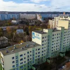 Duży blok mieszkalny S.M. Młyniec z charakterystycznym logo, otoczony innymi budynkami i drzewami jesienią.