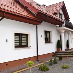 Biały dom z brązowym dachem i cokołem. Podjazd z kostki brukowej i kamyczków. Z czego zrobić cokół wokół domu?