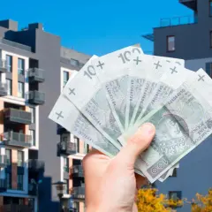 Dłoń trzyma banknoty 100 zł na tle nowoczesnych budynków mieszkalnych. Może to symbolizować podatek od sprzedaży mieszkania.