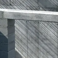 Betonowy nadproże nad otworem w budowie, podparty na bloczkach. Cienie ukośnie przecinają tło.