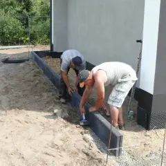 Dwóch mężczyzn montuje obrzeża. Używają młotka do osadzenia ich w suchym betonie, przygotowując teren pod przyszły ogród.