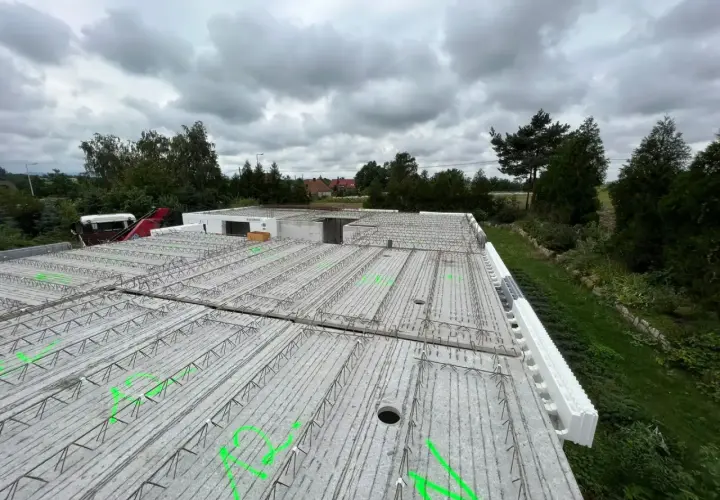 Budowa domu w toku. Widok na strop z prefabrykatów, zbrojenie czeka na beton. W tle drzewa i pochmurne niebo.