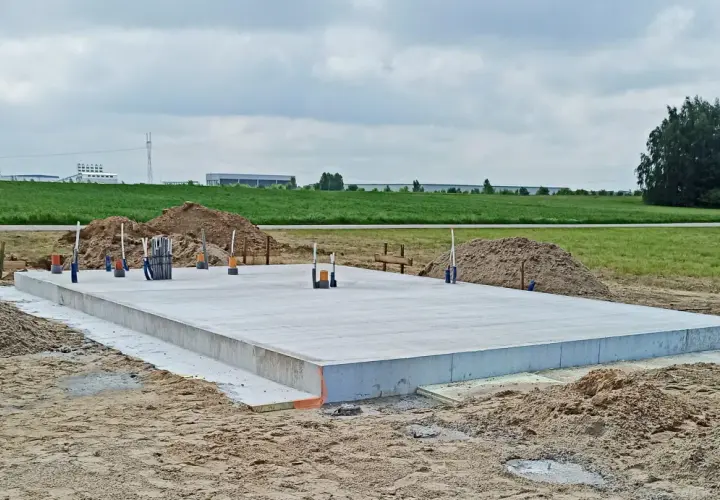 Gotowy projekt płyty fundamentowej na budowie. Betonowa płyta z wystającymi rurami i zbrojeniem, otoczona piaskiem i ziemią.