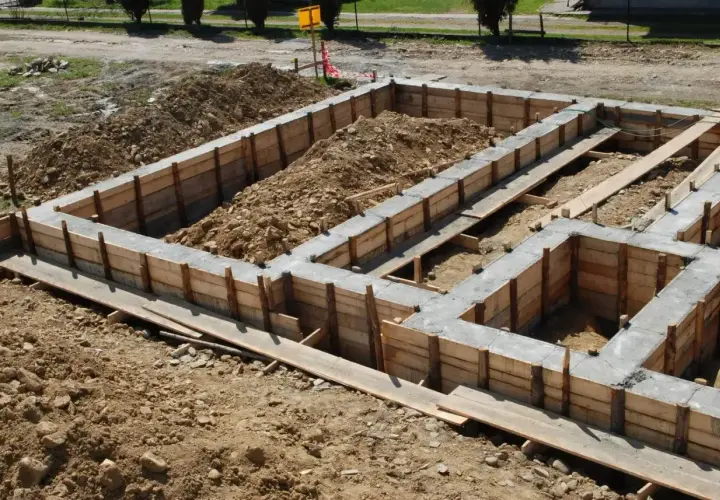 Budowa domu w toku: betonowe ławy fundamentowe w drewnianym szalunku, otoczone ziemią i gruzem.