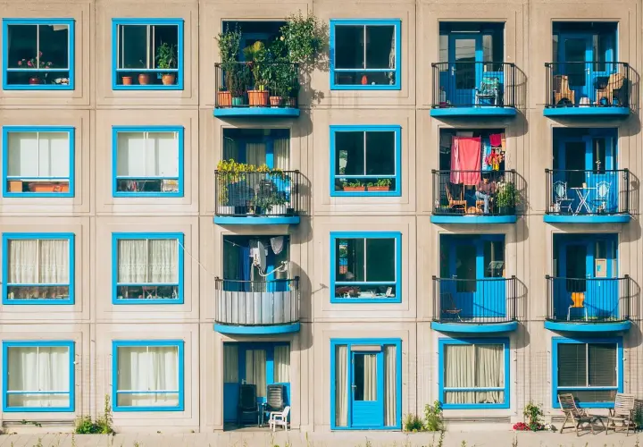 Wielopiętrowy budynek z balkonami, które są integralną częścią architektury, tworząc loggię. Niebieskie ramy okienne i drzwi dodają koloru.
