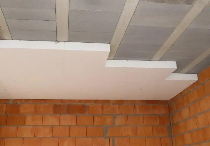 Ocieplenie stropu styropianem w trakcie budowy. Widoczne betonowe pustaki stropowe, płyty styropianowe i ściana z cegły.