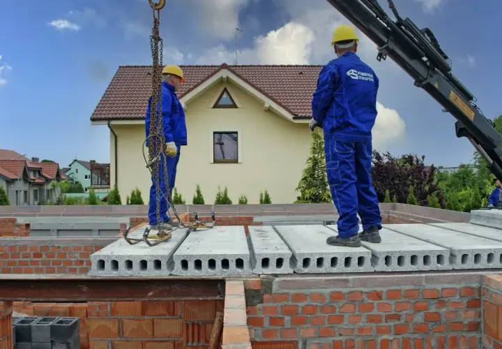 Dwóch pracowników w niebieskich kombinezonach montuje prefabrykowany strop. Dźwig podnosi kolejne płyty stropowe.