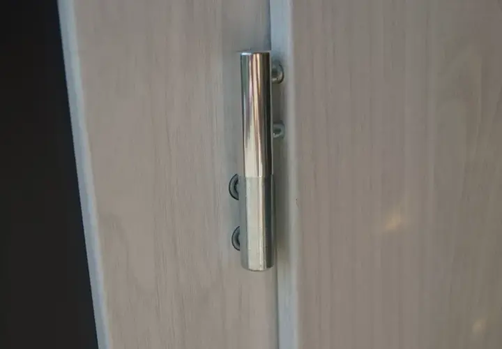 Metalowy zawias na jasnych drzwiach, umożliwiający regulację ich położenia.