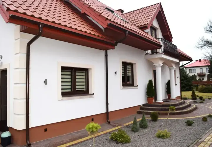 Biały dom z brązowym dachem i cokołem. Podjazd z kostki brukowej i kamyczków. Z czego zrobić cokół wokół domu?