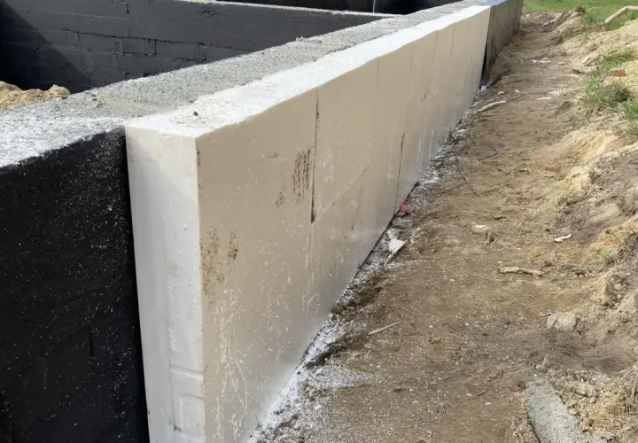 Izolowana podstawa pomnika z białego styropianu, przylegająca do czarnej, betonowej ściany fundamentowej.