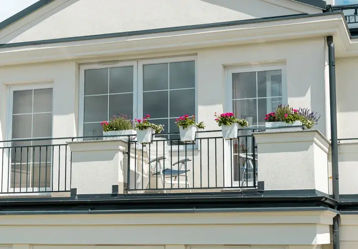 Białe okna na jasnej, teksturowanej elewacji budynku. Balkon z czarną balustradą i donicami z kwiatami.