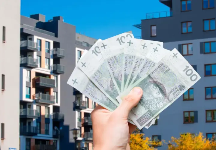 Dłoń trzyma banknoty 100 zł na tle nowoczesnych budynków mieszkalnych. Może to symbolizować podatek od sprzedaży mieszkania.
