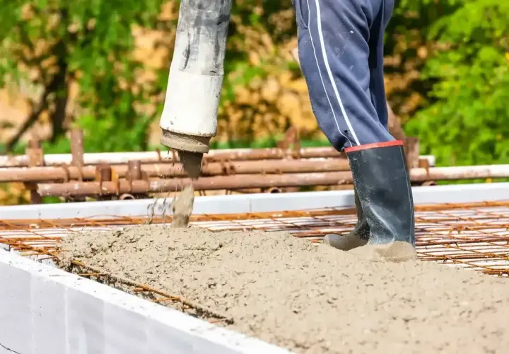 Beton wylewany jest na zbrojenie w szalunku. Pracownik w gumowcach stoi w świeżym betonie.