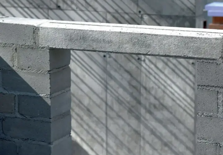 Betonowy nadproże nad otworem w budowie, podparty na bloczkach. Cienie ukośnie przecinają tło.