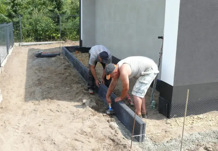 Dwóch mężczyzn montuje obrzeża. Używają młotka do osadzenia ich w suchym betonie, przygotowując teren pod przyszły ogród.