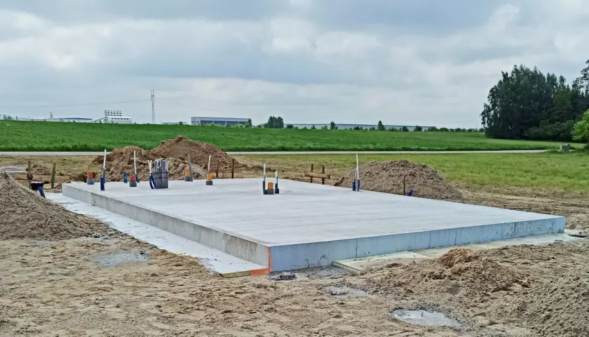 Gotowy projekt płyty fundamentowej na budowie. Betonowa płyta z wystającymi rurami i zbrojeniem, otoczona piaskiem i ziemią.