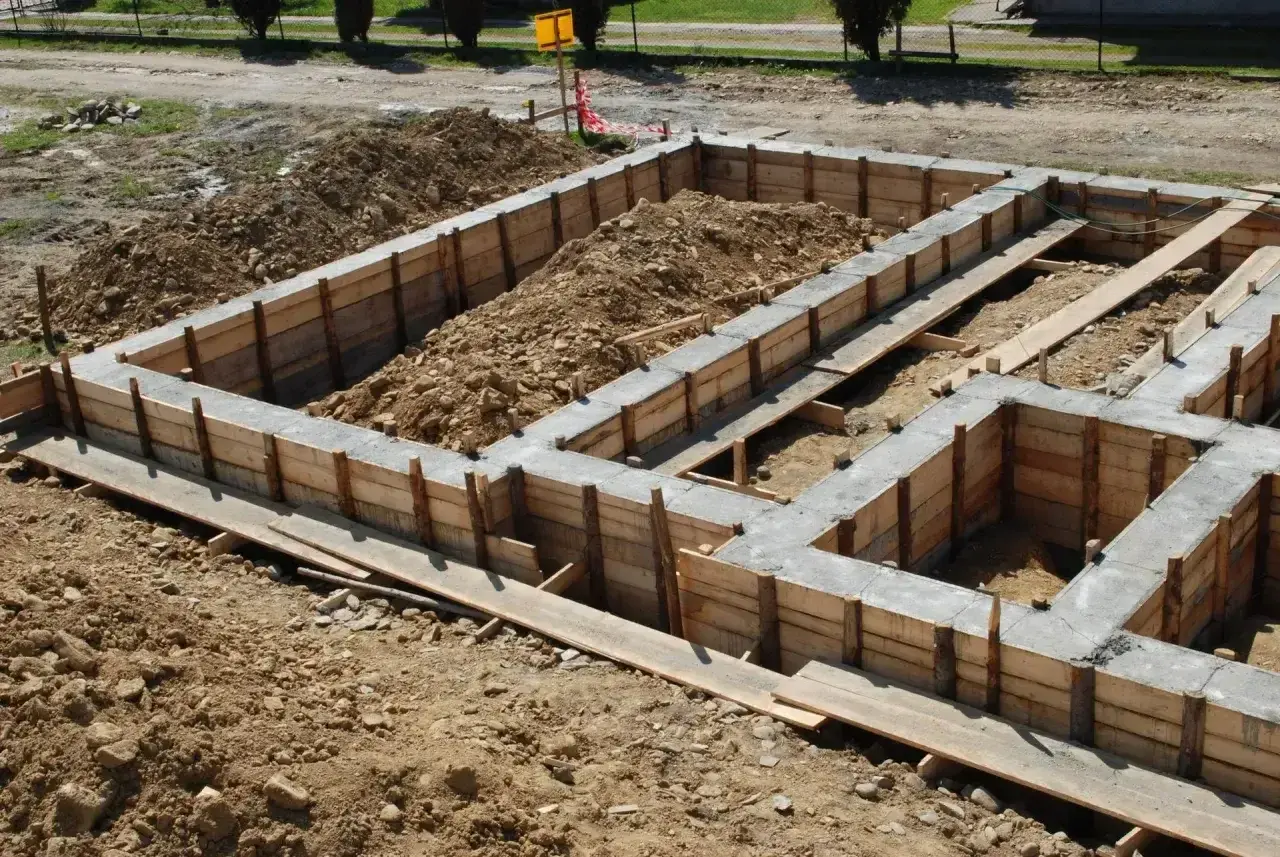 Budowa domu w toku: betonowe ławy fundamentowe w drewnianym szalunku, otoczone ziemią i gruzem.