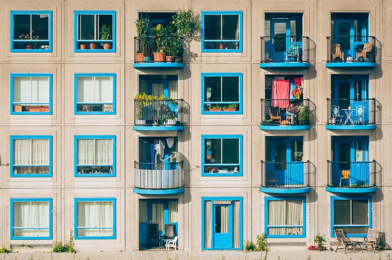 Wielopiętrowy budynek z balkonami, które są integralną częścią architektury, tworząc loggię. Niebieskie ramy okienne i drzwi dodają koloru.