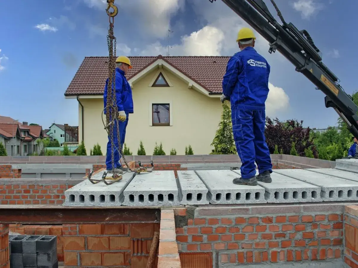 Dwóch pracowników w niebieskich kombinezonach montuje prefabrykowany strop. Dźwig podnosi kolejne płyty stropowe.