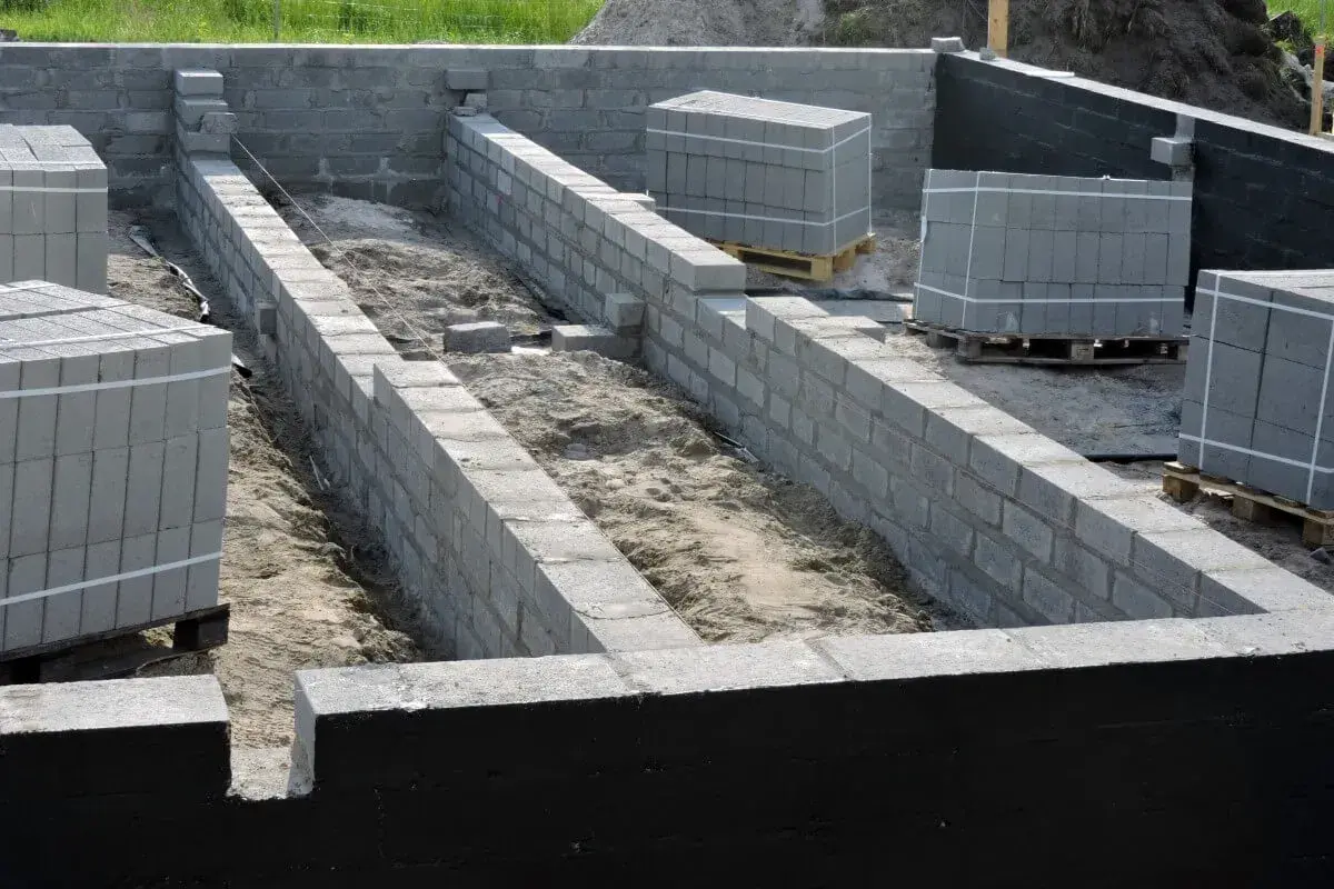Budowa fundamentów z bloczków betonowych. Widać ułożone palety z bloczkami i rozpoczęte mury. Folia fundamentowa zabezpiecza konstrukcję.