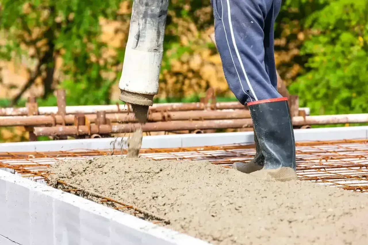 Beton wylewany jest na zbrojenie w szalunku. Pracownik w gumowcach stoi w świeżym betonie.