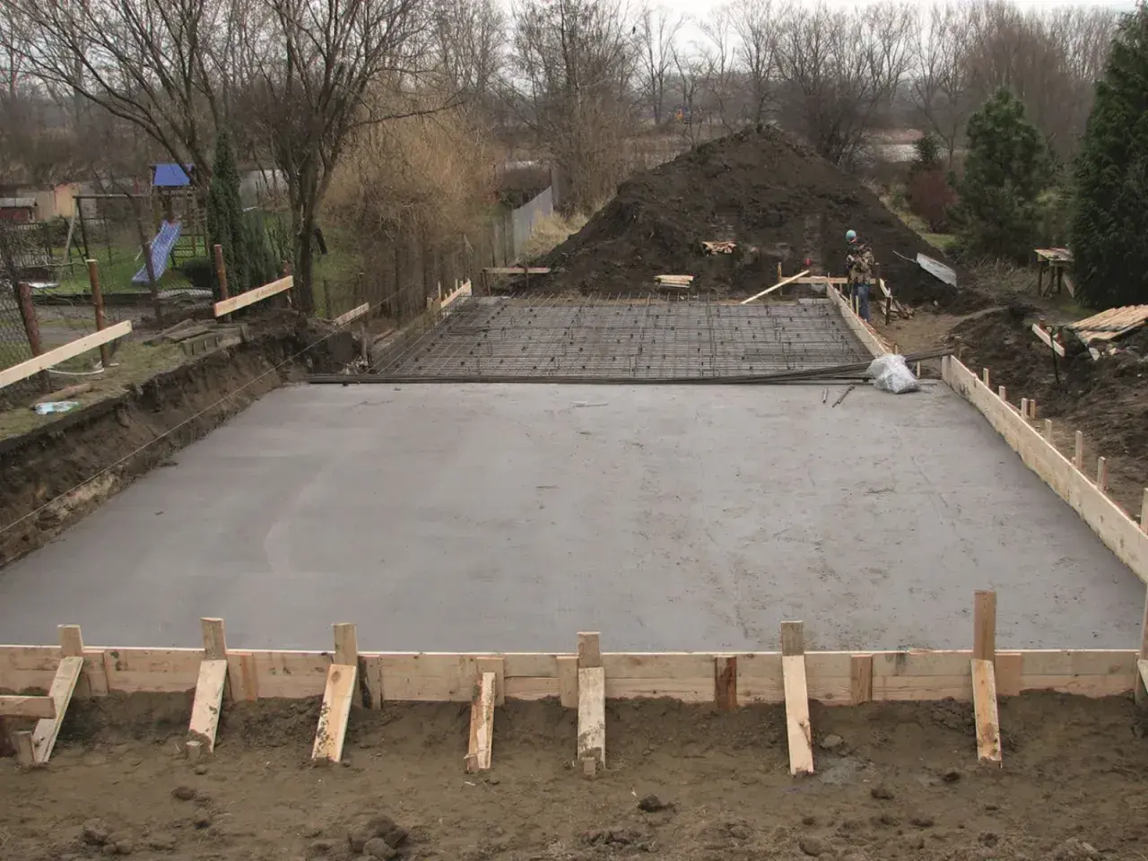 Plac budowy z wylana płytą fundamentową, przygotowaną pod klasę ekspozycji betonu. Widać zbrojenie i szalunki.