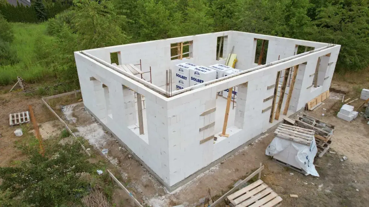 Budowa domu z bloczków z betonu komórkowego. Na górze widoczne belki stropowe, a na dole palety z materiałami budowlanymi. Uważaj na mostek termiczny przy oknach.