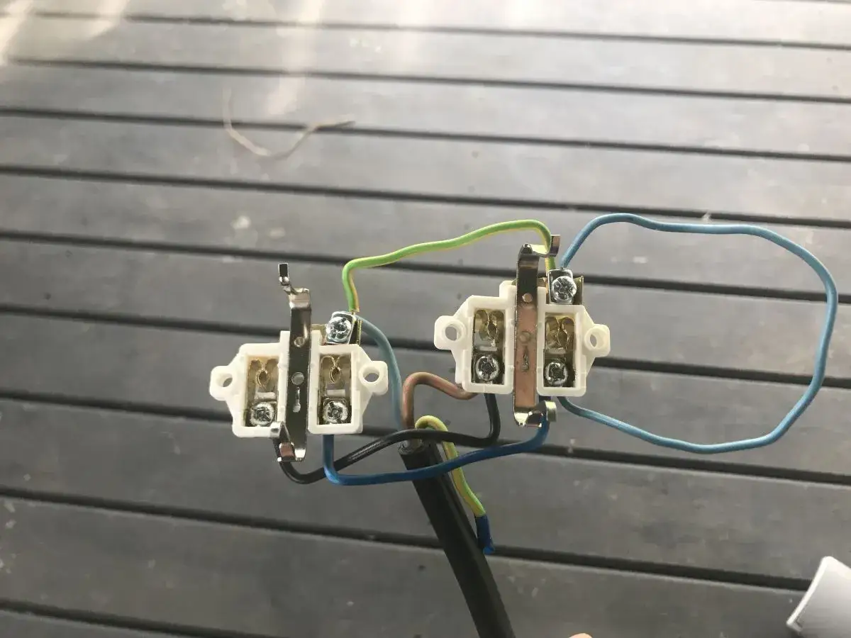 Mostkowanie podwójnego gniazdka elektrycznego. Widać białe obudowy, metalowe styki i kolorowe przewody: zielony, niebieski i brązowy.
