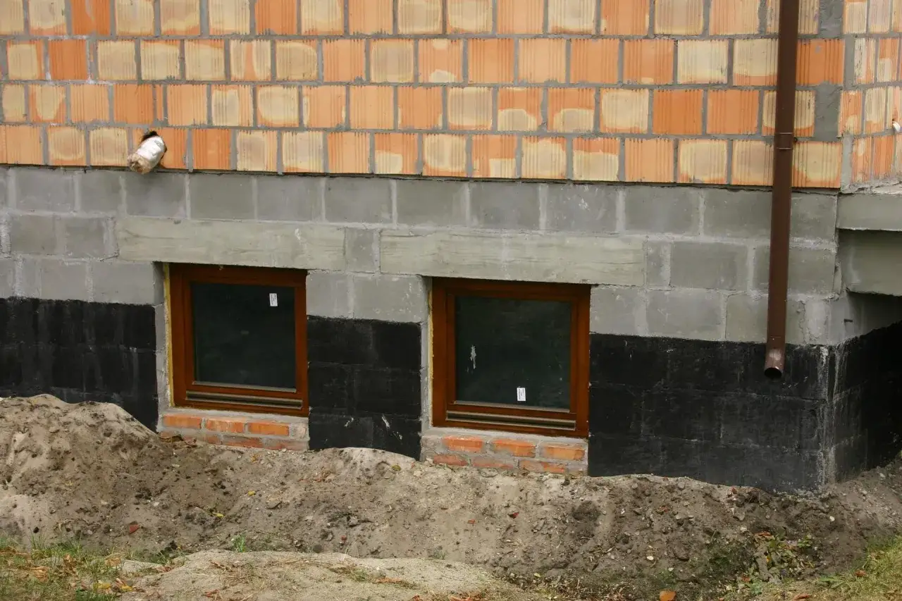 Budowa domu w toku. Widoczne okna piwniczne, izolacja fundamentów i ściany z pustaków. Trwa podbijanie fundamentów.
