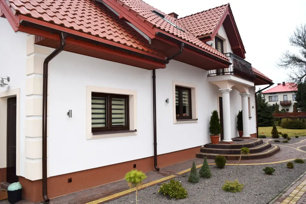 Biały dom z brązowym dachem i rynnami. Elewacja ozdobiona kamiennymi elementami, a u podstawy widoczny brązowy cokół.