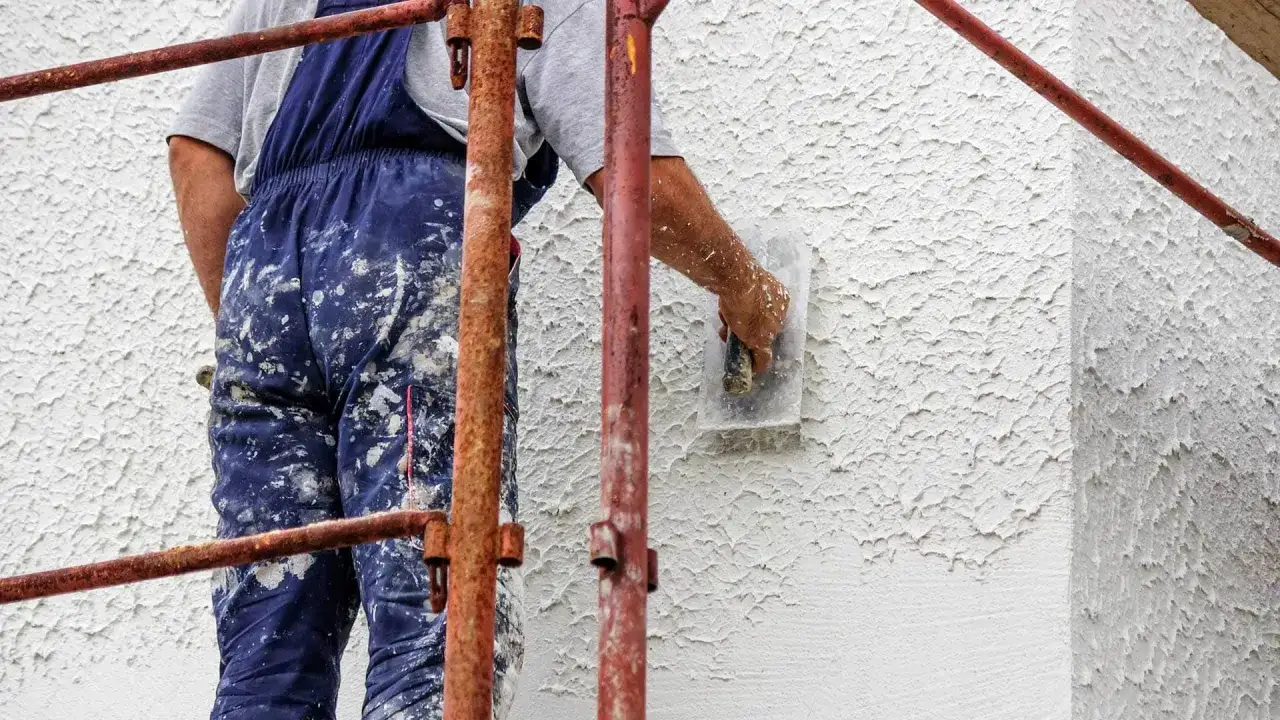 Pracownik w niebieskim kombinezonie nakłada tynki mineralne na ścianę za pomocą kielni.